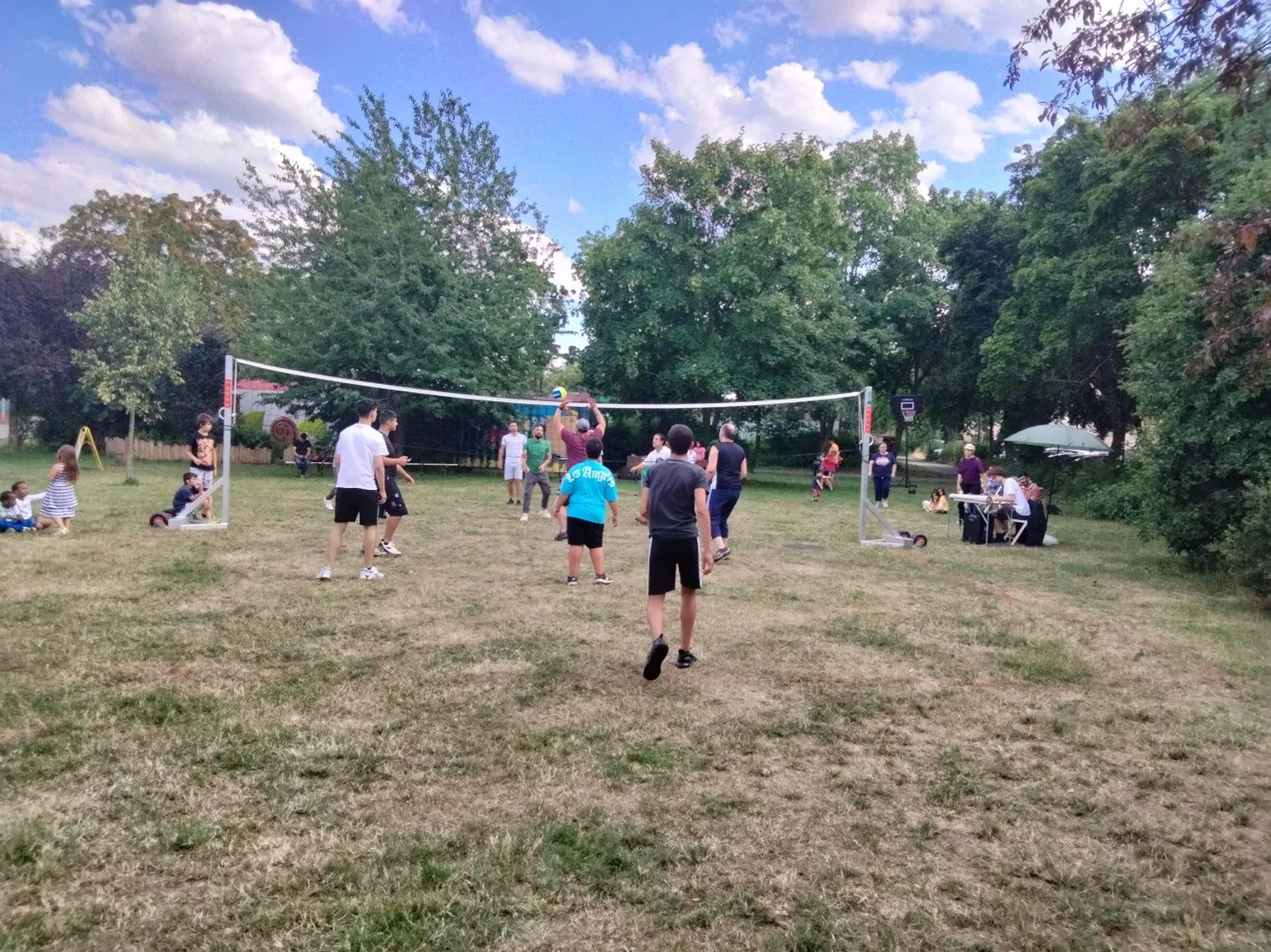 Es ist ein Park zusehen, in dem ein Volleyballnetz aufgebaut ist. Es vielen etwa 12 Personen auf dem Bild zusammen Volleyball.