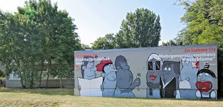 Auf dem Haus ist ein Graffiti gesprüht. Das Graffiti zeigt 7 Personen, die unterschiedliche Sachen machen. Eine Person liest Zeitung, eine andere ein Buch, eine pustet eine Pusteblume, eine hat einen Schnuller im Mund und eine eine große Sonnenbrille auf. Links steht: Zur Saaleaue 25a Stadtteilbibliothek West. Rechts steht Zur Saaleaue 51a Mehrgenerationenhaus Pusteblume. Die beiden Orte liegen nebeneinander. 