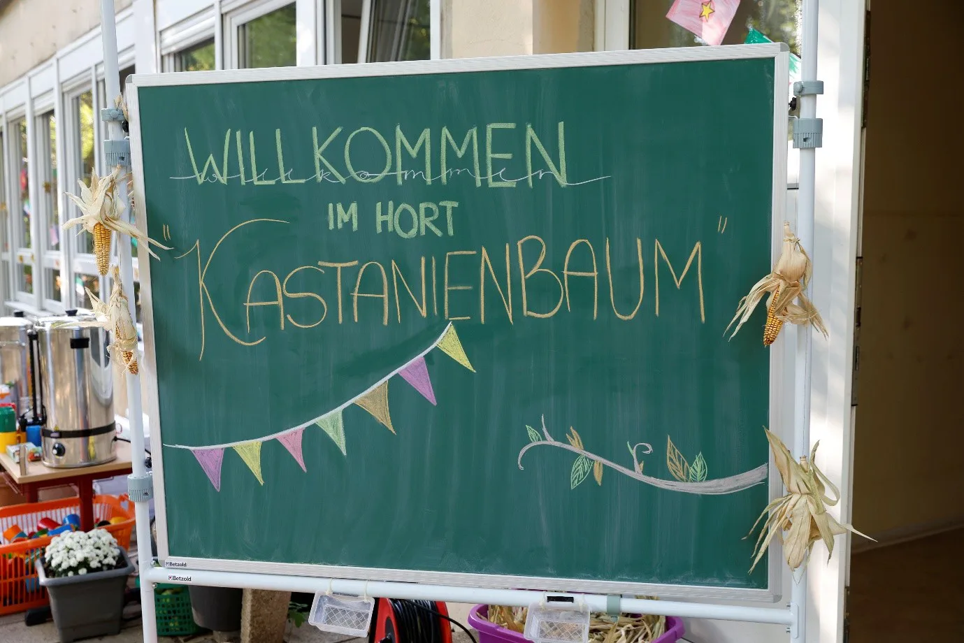 Auf einer grünen Schultafel steht in bunten Buchstaben Willkommen im Hort Kastanienbaum. Darunter ist ein Zweig und eine Girlande gemalt.